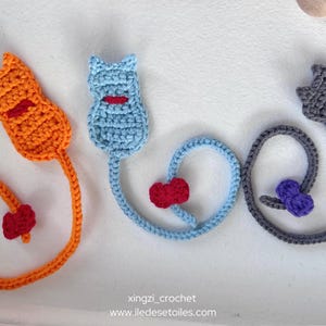 May include: Three crocheted cat tail bookmarks in different coloured yarn. The orange cat has a red bow, the blue cat has a red bow, and the grey cat has a purple bow. The tails are curled into a heart shape.