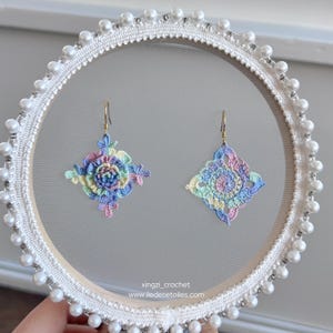 May include: A pair of crocheted earrings with a rainbow colour scheme. The earrings are displayed on a white circular frame with pearl accents. The text "xingzi_crochet www.iledesetoiles.com" is visible at the bottom of the image.