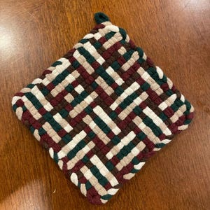 May include: A woven pot holder with a square shape and a loop handle. The pot holder is made of a thick, textured fabric in shades of brown, green, white, and burgundy.