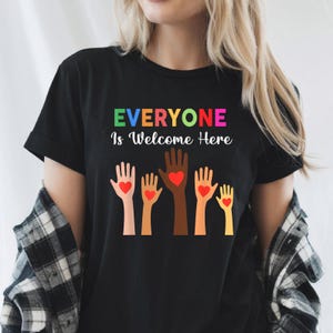 以下が含まれることがあります： レインボーカラーで「Everyone is Welcome Here」と書かれた黒いTシャツ。歓迎のしるしとして、ハートを持った5本の腕が突き上げられている。