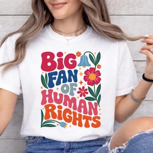May include: White t-shirt with a colorful graphic that reads "Big Fan of Human Rights." The design features retro-style lettering and floral elements in red, blue, orange, and green. A casual, comfortable tee.