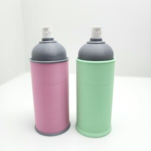 Puede incluir: Dos botes de pintura en aerosol coloridos. Uno es rosa, el otro verde. Ambos tienen tapas grises y boquillas blancas. Los botes son cilíndricos y están sobre una superficie blanca.