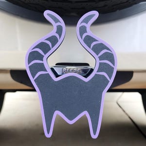 May include: A black and purple trailer hitch cover featuring a stylized design with curved horns. The hitch cover is mounted on a vehicle, with the brand name "REESE" visible. The design is outlined in purple.