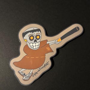 Puede incluir: Una pegatina con un esqueleto de dibujos animados con una túnica marrón, sosteniendo un bate de béisbol. El esqueleto tiene un cráneo decorado con detalles en naranja y amarillo. El logotipo de los Padres de San Diego está en la túnica. La firma del artista es visible.