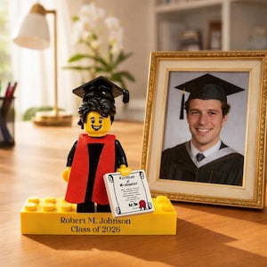 May include: A Lego graduation figure wearing a black mortarboard and red academic regalia, holding a diploma. The figure stands on a yellow base with the name "Robert M. Johnson Class of 2026". A framed graduation photo is next to the figure.