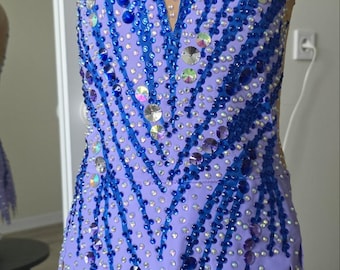 Peut inclure: Un justaucorps sans manches avec une base violet clair et un haut en maille couleur chair. Le vêtement est richement orné de cristaux bleus et irisés disposés en motifs verticaux en forme de V sur le devant et l'ourlet dentelé.