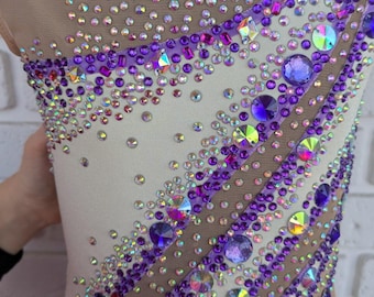 Peut inclure: Justaucorps crème avec dos et manches en résille nude. Il est orné de strass violets et irisés disposés en rayures diagonales et motifs épars, créant un effet scintillant, idéal pour la danse ou la gymnastique.