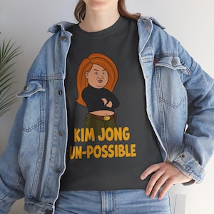 May include: Dark gray t-shirt featuring a cartoon illustration of a woman with red hair and the text "KIM JONG UN-POSSIBLE" in yellow. The woman is wearing a black top and olive green pants. The shirt is paired with a denim jacket.