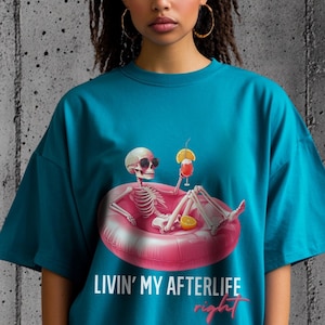 Op de afbeelding: Tealkleurig T-shirt met een afbeelding van een skelet met zonnebril, liggend in een roze opblaasbare ring en een cocktail vasthoudend. De tekst "Livin' My Afterlife right" staat eronder. Het shirt heeft een relaxte, oversized pasvorm.