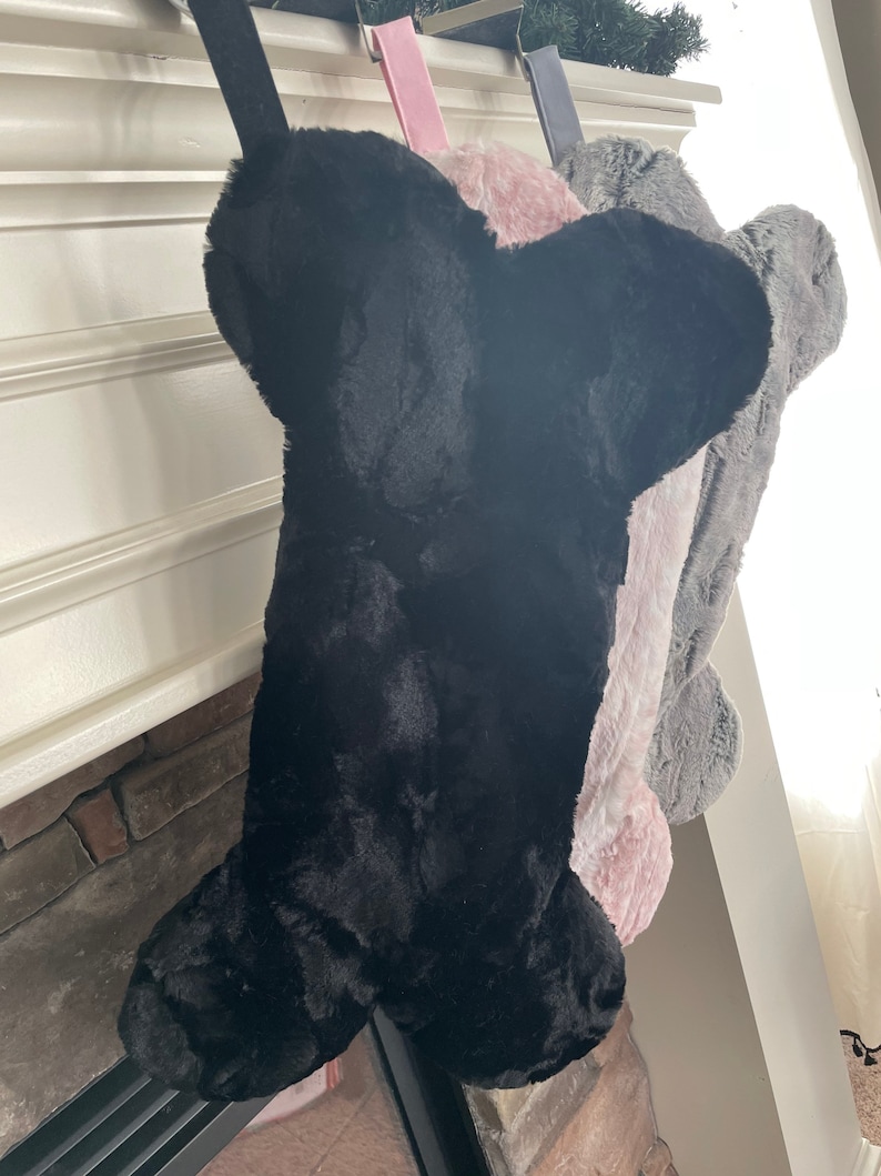 May include: Three plush dog bone stockings hanging from a fireplace mantel. The stockings are black, pink, and gray.