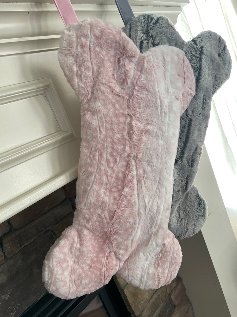 May include: Two plush dog bone stockings, one pink and one gray, hanging from a fireplace mantel.