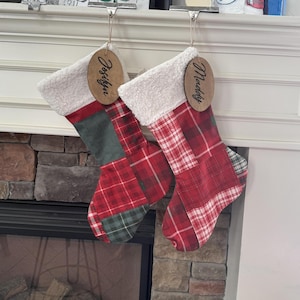 May include: Two Christmas stockings with red and green plaid patterns and white faux fur cuffs hang from a fireplace mantel. Each stocking has a wooden tag with a name written on it. The stockings are a festive holiday decoration.