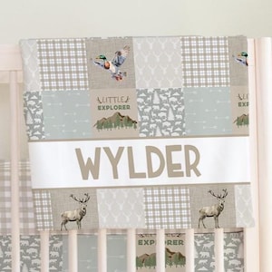 May include: A patchwork baby blanket with a woodland theme. The blanket features squares with deer, ducks, trees, and plaid patterns in shades of gray, tan, and white. The name "WYLDER" is displayed in large, tan letters across the center.