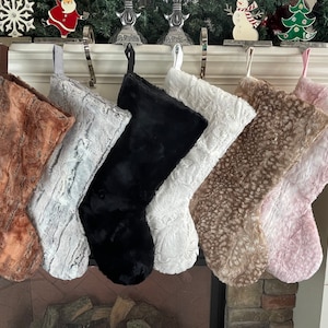 May include: A collection of seven plush Christmas stockings in various colors, including brown, gray, black, white, and pink. Each stocking is made of a soft, textured material and hangs from a decorative mantelpiece.
