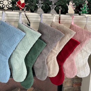 May include: A collection of seven Christmas stockings in various colors, including blue, green, gray, red, and pink. Each stocking has a diamond pattern and a loop for hanging. The stockings are displayed on a fireplace mantel.