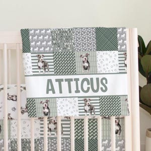 May include: A crib bumper with a patchwork design in shades of gray and green. The bumper features images of dogs, paw prints, and bones. The name "ATTICUS" is displayed in large, bold letters across the center.