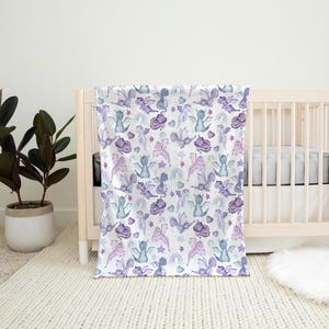 May include: A white baby blanket with a purple and blue watercolor pattern of dragons and stars. The blanket is draped over a white crib with a white mattress.
