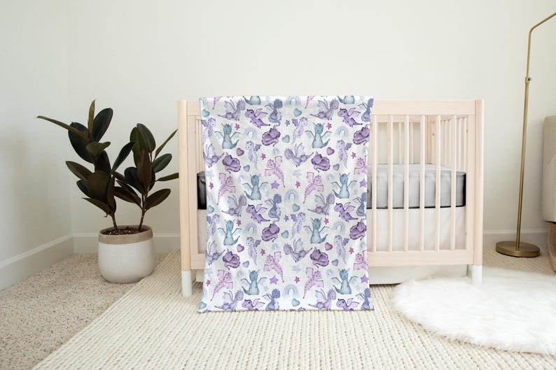 May include: A white baby blanket with a purple and blue watercolor pattern of dragons and stars. The blanket is draped over a white crib with a white mattress.