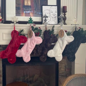 May include: Five plush dog bone-shaped Christmas stockings hanging on a fireplace mantel. The stockings are red, pink, brown, white, and black. Each stocking has a personalized wooden tag with a name on it. The names are Megan, Elizabeth, and two others that are not visible.