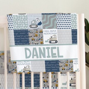 May include: A cot blanket with a patchwork design featuring construction vehicles in shades of grey, blue, and yellow. The blanket has the name "DANIEL" and the phrase "I WILL MOVE MOUNTAINS".