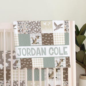 May include: A white crib bumper with a patchwork design featuring brown, green, and white ducks. The bumper has a personalized name banner that reads "JORDAN COLE".