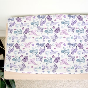 May include: A white changing pad cover with a purple and blue dragon pattern. The pattern includes stars, hearts, and rainbows.