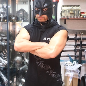 Puede incluir: Una persona con una máscara de Batman negra y una camiseta negra sin mangas con un estampado de camuflaje gris. La camiseta tiene la palabra "BUTZ" impresa. La persona está de pie frente a una pantalla de cascos y espadas.
