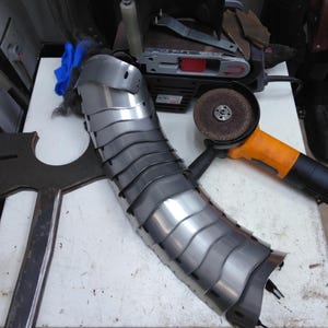 May include: A silver metal armour piece with a curved shape, likely for a forearm or elbow. The armour is made of multiple overlapping plates. The piece is lying on a white surface next to a black and orange power tool.
