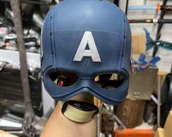 May include: A navy blue Captain America helmet with a silver "A" emblem. The helmet has eye cutouts and a chin strap. The image is taken from a slightly low angle, showcasing the back of the helmet.