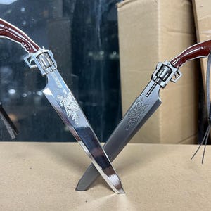 May include: Two silver-colored pocket knives with red handles, crossed in an X shape. The knives have intricate designs on the blades and are attached to black leather cords.