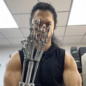 Könnte beinhalten: Eine Person mit langen schwarzen Haaren hält eine silberne Roboterhand vor ihrem Gesicht. Die Hand ist voll beweglich und hat ein realistisches Aussehen.