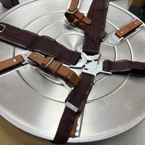 May include: A silver metal shield with a brown leather strap and a dark brown fabric strap. The shield has a circular shape and is attached to the straps with metal buckles.