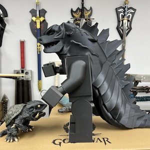 May include: A black Godzilla Lego figure stands next to a smaller black turtle monster figure. Both figures are made of plastic and have a detailed design.