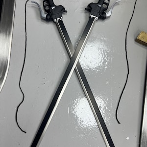 Könnte beinhalten: Zwei schwarze und silberne Schwerter mit schwarzen Griffen und Schnüren, die an den Griffen befestigt sind. Die Schwerter sind gekreuzt und die Schnüre hängen herunter.