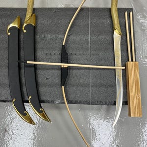 May include: A fantasy set comprising two swords in black sheaths with gold detailing, a wooden bow, arrows, and a wooden arrow holder. The swords have gold handles and etched blades. The bow is made of light-coloured wood.