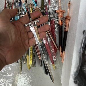 May include: A collection of miniature swords with various finishes, including silver, gold, and red. The swords are displayed in a hand, with the tips pointing upwards.