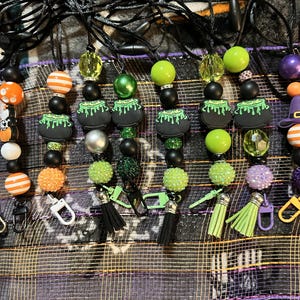 May include: Several beaded lanyards with Halloween-themed charms. The lanyards feature black beads, green and orange accents, and charms like witch cauldrons and orange striped beads. Each lanyard has a clasp and a black tassel.