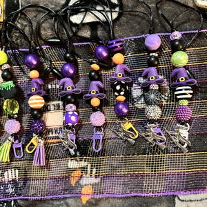 以下が含まれることがあります： ハロウィーンをテーマにしたビーズのランヤードのコレクション。各ランヤードは、紫、オレンジ、黒、緑のさまざまなビーズが付いた黒いコードが特徴です。チャームには、魔女の帽子、大釜、ほうきが含まれます。ランヤードにはロブスタークラスプが付いています。