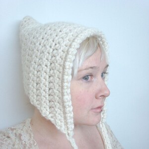 May include: A cream-colored, chunky knit bonnet featuring a textured, ribbed stitch pattern. The headwear has a pointed top, a rounded face opening, and two long, braided ties extending from the bottom edges to secure under the chin.