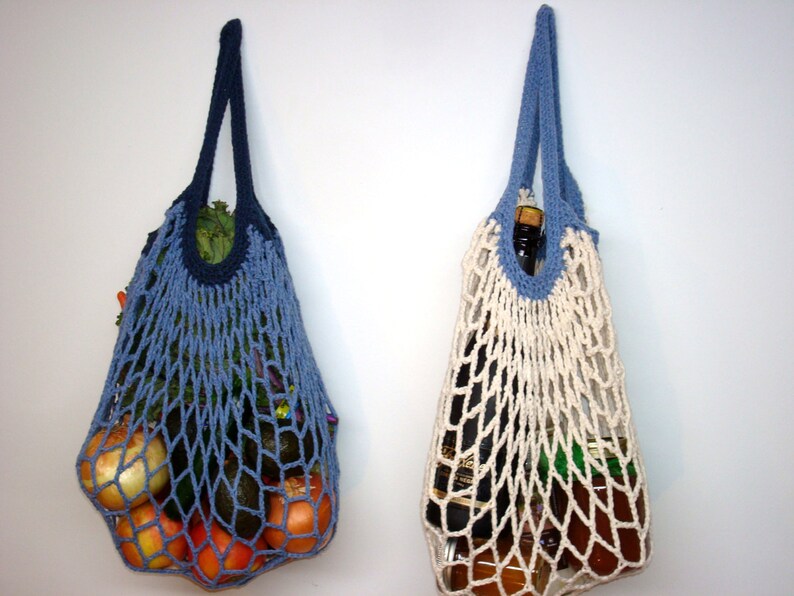 May include: Two crocheted string bags, one blue and one white, hanging on a white wall. The blue bag is filled with produce, including onions, apples, and avocados. The white bag is filled with a glass bottle and jars.