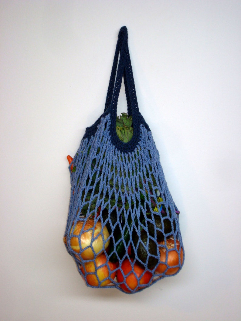 May include: A blue crocheted mesh bag filled with produce, including oranges, avocados, and onions. The bag is hanging on a white wall.