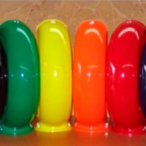 Puede incluir: Una colección de ocho objetos circulares, huecos y con forma de anillo dispuestos en fila. Los artículos tienen un acabado brillante de color sólido, incluyendo blanco, negro, verde, amarillo, naranja, rojo, azul oscuro y azul brillante, cada uno con una pequeña base.