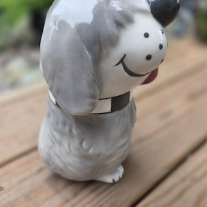 Op de afbeelding: Een keramieken hondenbeeldje met een glanzende grijze afwerking, een wit gezicht met zwarte stippen en een zwarte neus. De hond heeft een zwart-witte halsband om de nek en een geschilderde zwarte glimlach. Het figuur heeft textuur in de vacht.