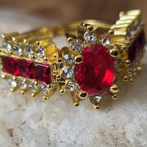 May include: A gold-toned ring with a large, oval-cut red gemstone at the center, flanked by smaller red and clear stones. The ring has a detailed, ornate design with a vintage aesthetic, suitable for special occasions.