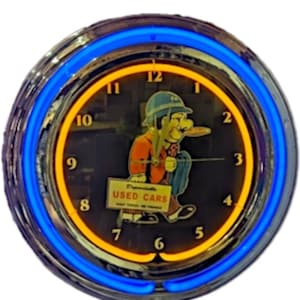 Op de afbeelding: Een ronde wandklok met een chromen frame en een helderblauwe neonring. De wijzerplaat toont een stripfiguur met een hoed en oranje jas die een bord vasthoudt met de tekst "Dependable USED CARS" en "EASY TERMS - WE FINANCE" in zwarte letters.