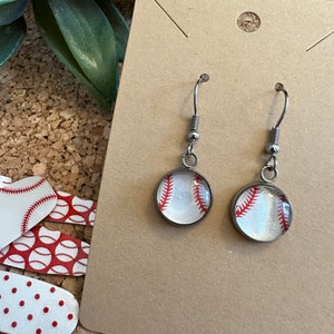 May include: A pair of silver baseball earrings with white and red baseballs. The earrings are on a brown card.