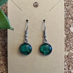 May include: A pair of silver dangle earrings with green round resin cabochons. The cabochons have a sparkly, glittery effect.