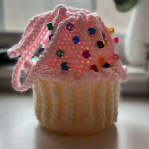 Peut inclure: Un coussin &agrave; &eacute;pingles en forme de cupcake crochet&eacute; en rose et blanc avec des perles color&eacute;es pour la d&eacute;coration.