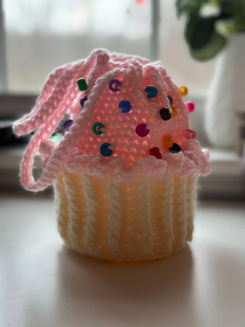 Peut inclure: Un coussin &agrave; &eacute;pingles en forme de cupcake crochet&eacute; en rose et blanc avec des perles color&eacute;es pour la d&eacute;coration.