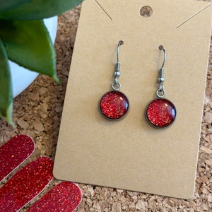 May include: A pair of silver dangle earrings with red glitter round charms. The earrings are displayed on a brown card.